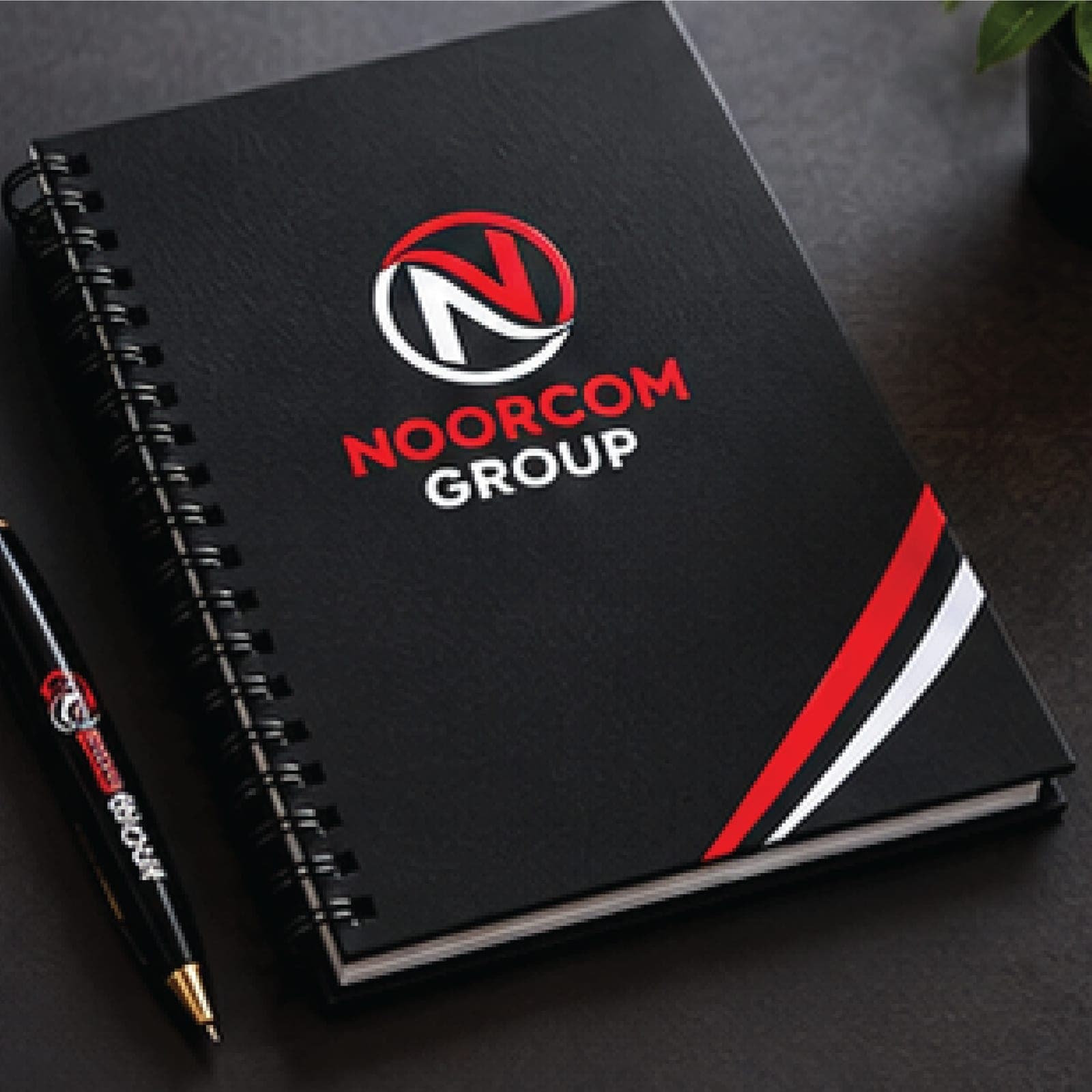 A black spiral notebook with the Noorcom Group logo and a branded pen